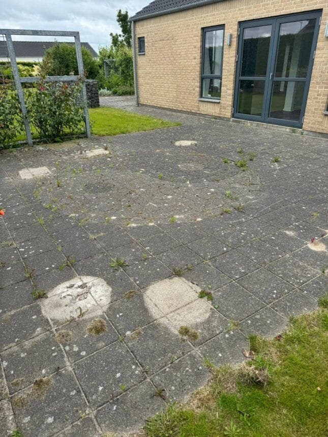 Flisebelagt terrasse med ukrudt ved murstenshus